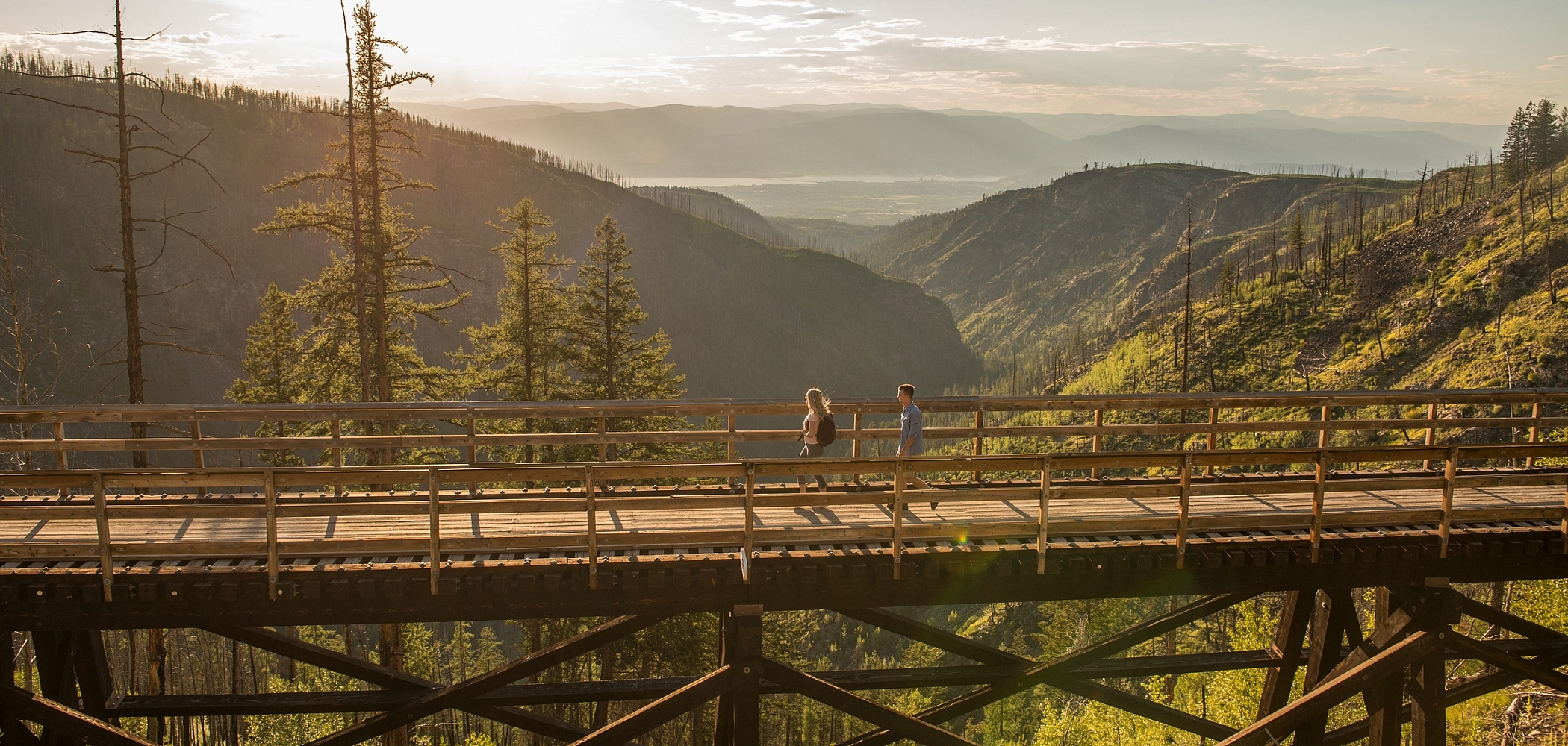 Two people walk across a tall wooden trestle bridge with forested mountains in the background.