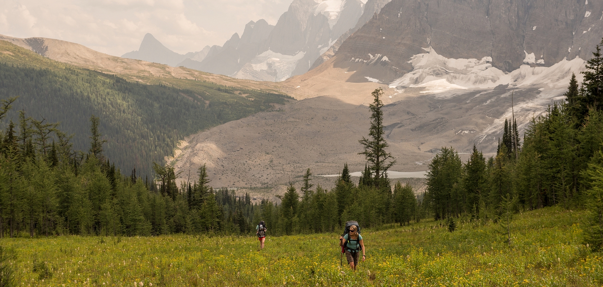 Two people with big backpacks walk toward the camera through a grassy field. Behind them is a ring of evergreen trees, and beyond those are rugged mountain peaks and glaciers.