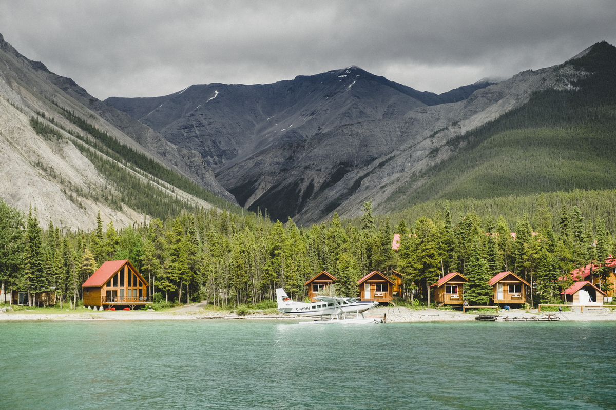 The rustic cabins of Northern Rockies Lodge perched on the turqoise lakeshore of Muncho Lake with mountains in the background and a float plane in the foreground.