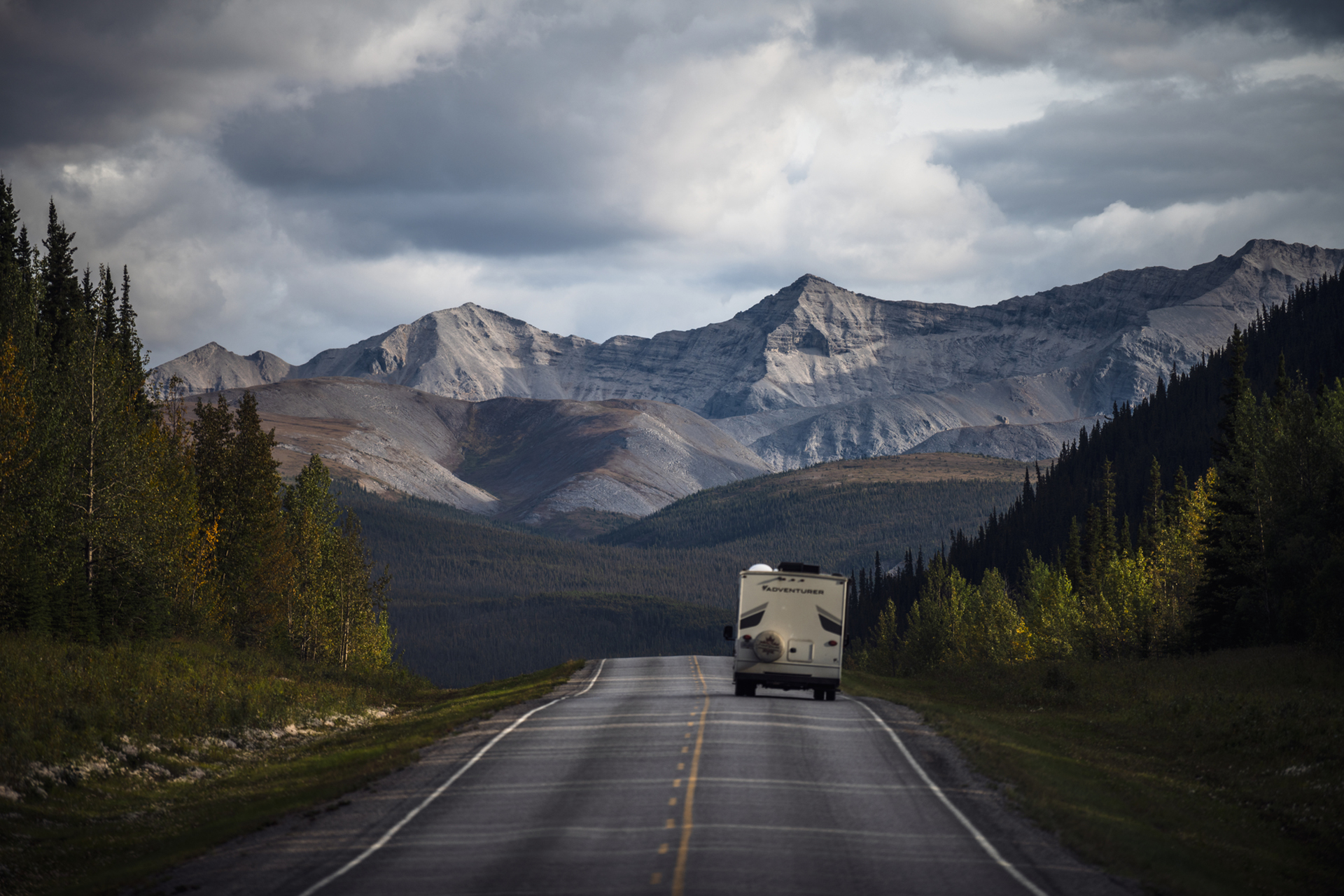 An RV drives down a quiet highway towards a mountain range in the distance. Words on the back of the RV read, "Adventures."